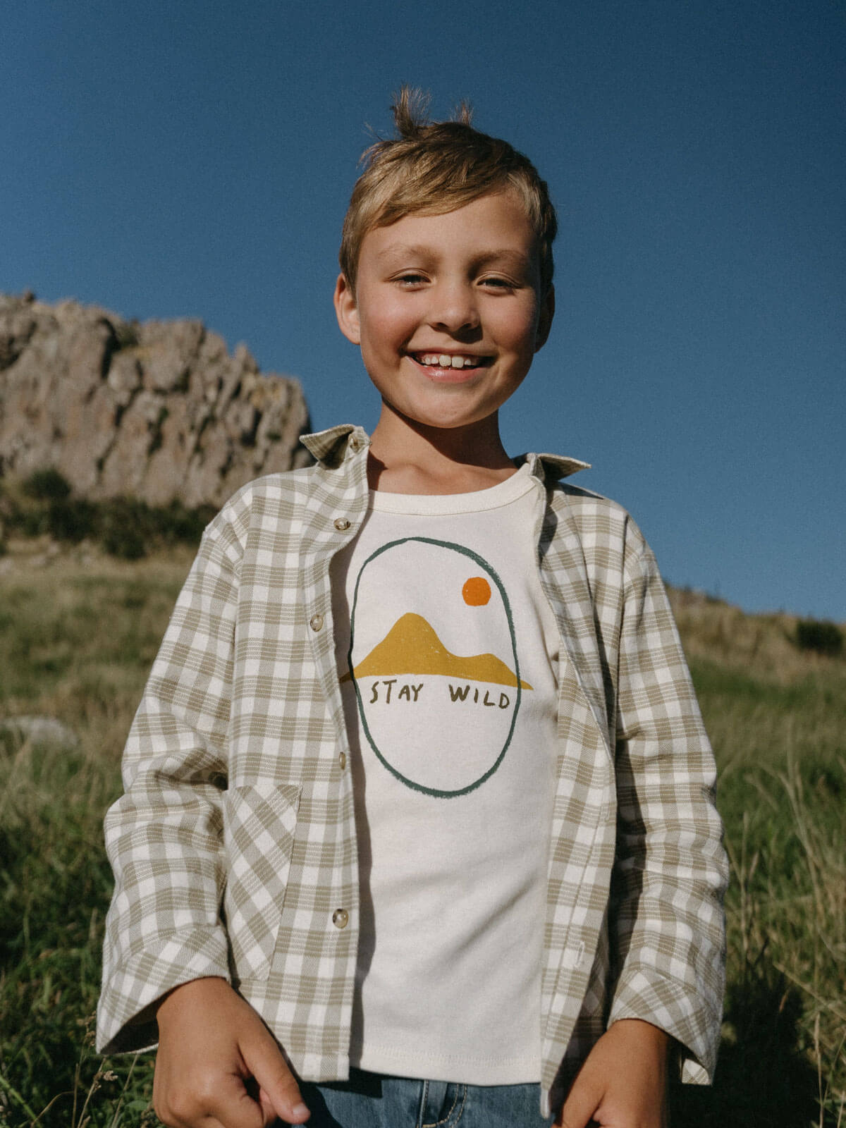 Child wearing a 'Stay Wild' t-shirt in a natural setting with mountains.