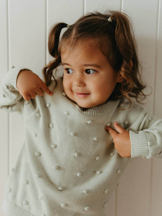 Child wearing a light green sweater with white polka dots against a white wall.