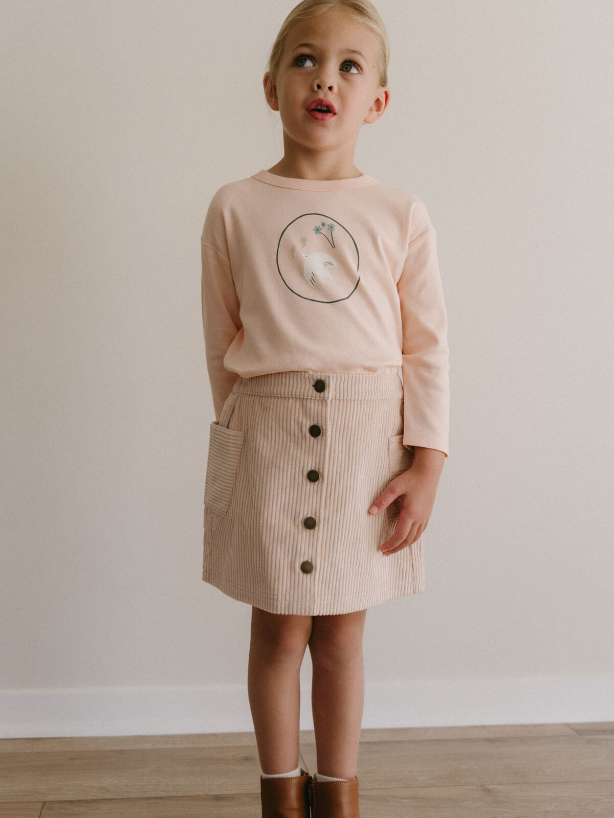 Young girl wearing a pink sweater with a graphic and a beige buttoned skirt against a plain background
