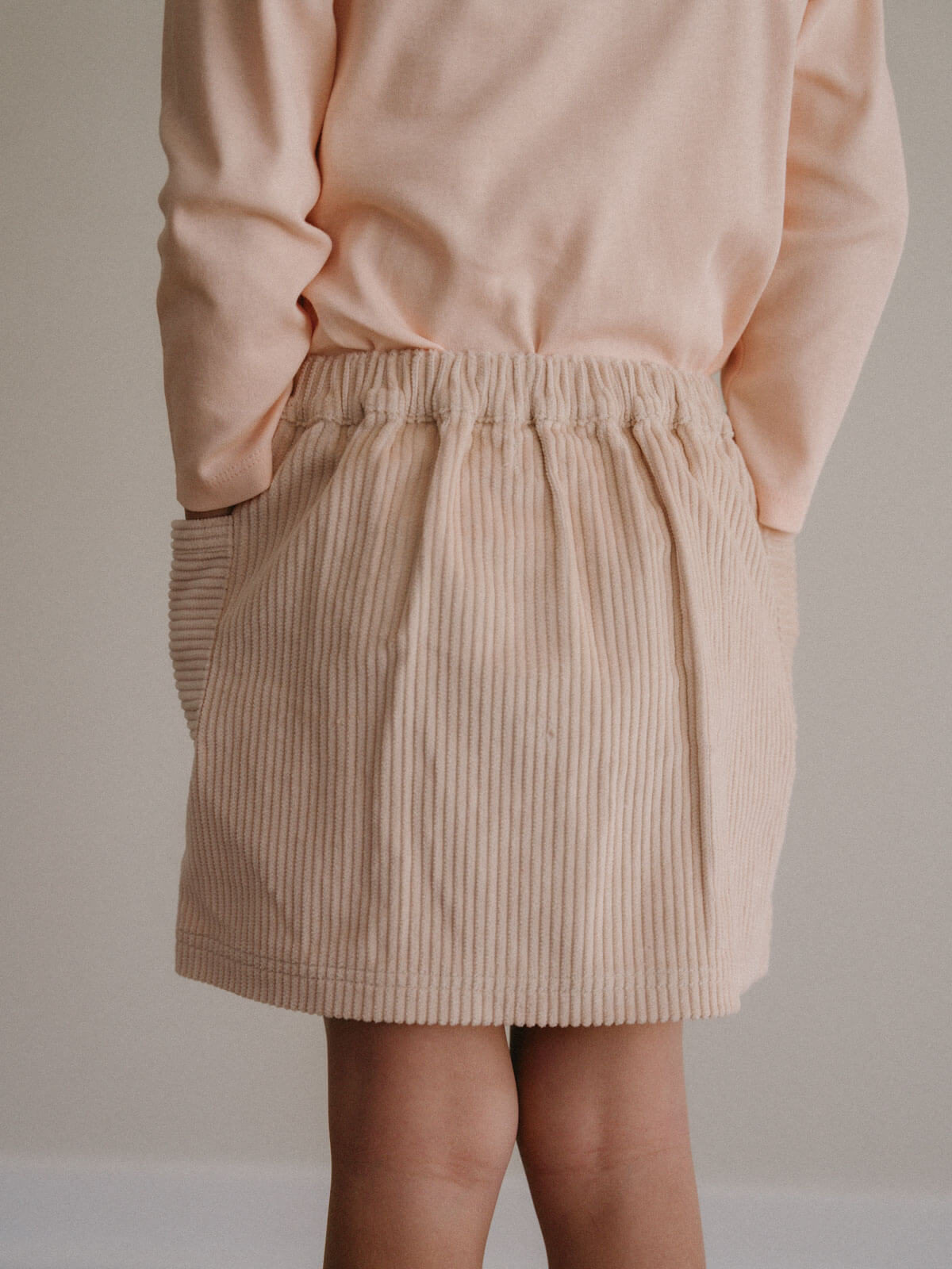 girl wearing a beige corduroy skirt and peach long-sleeve top against a neutral background