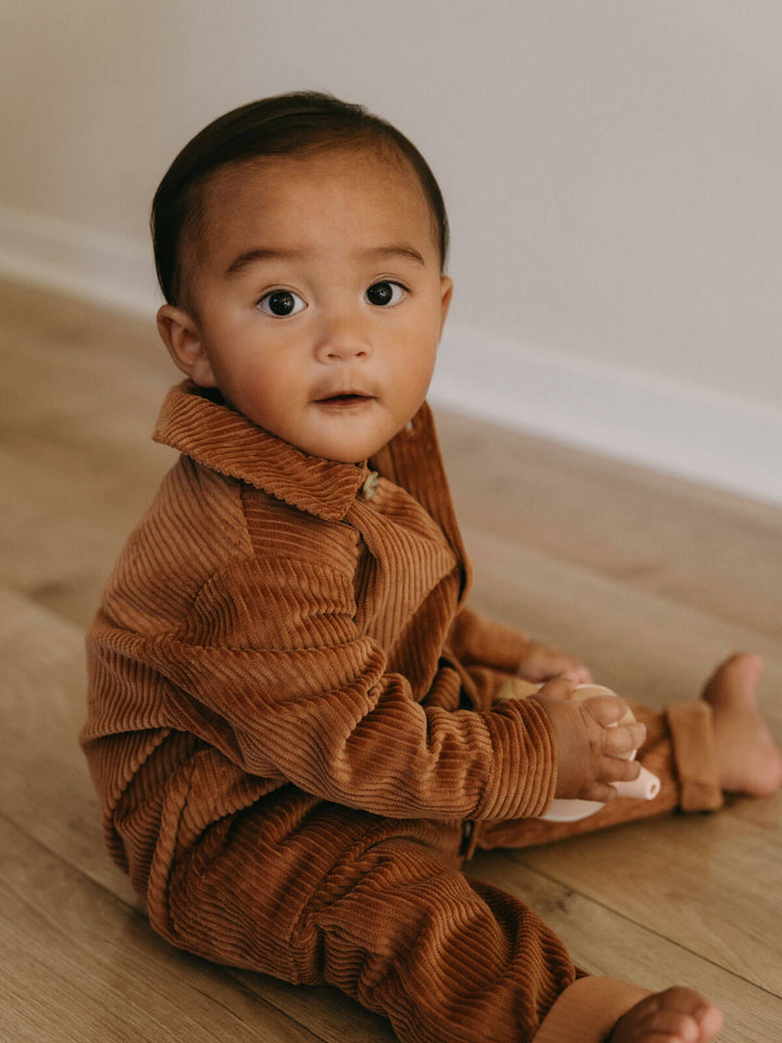 Baby wearing a brown corduroy outfit sitting on a wooden floor.