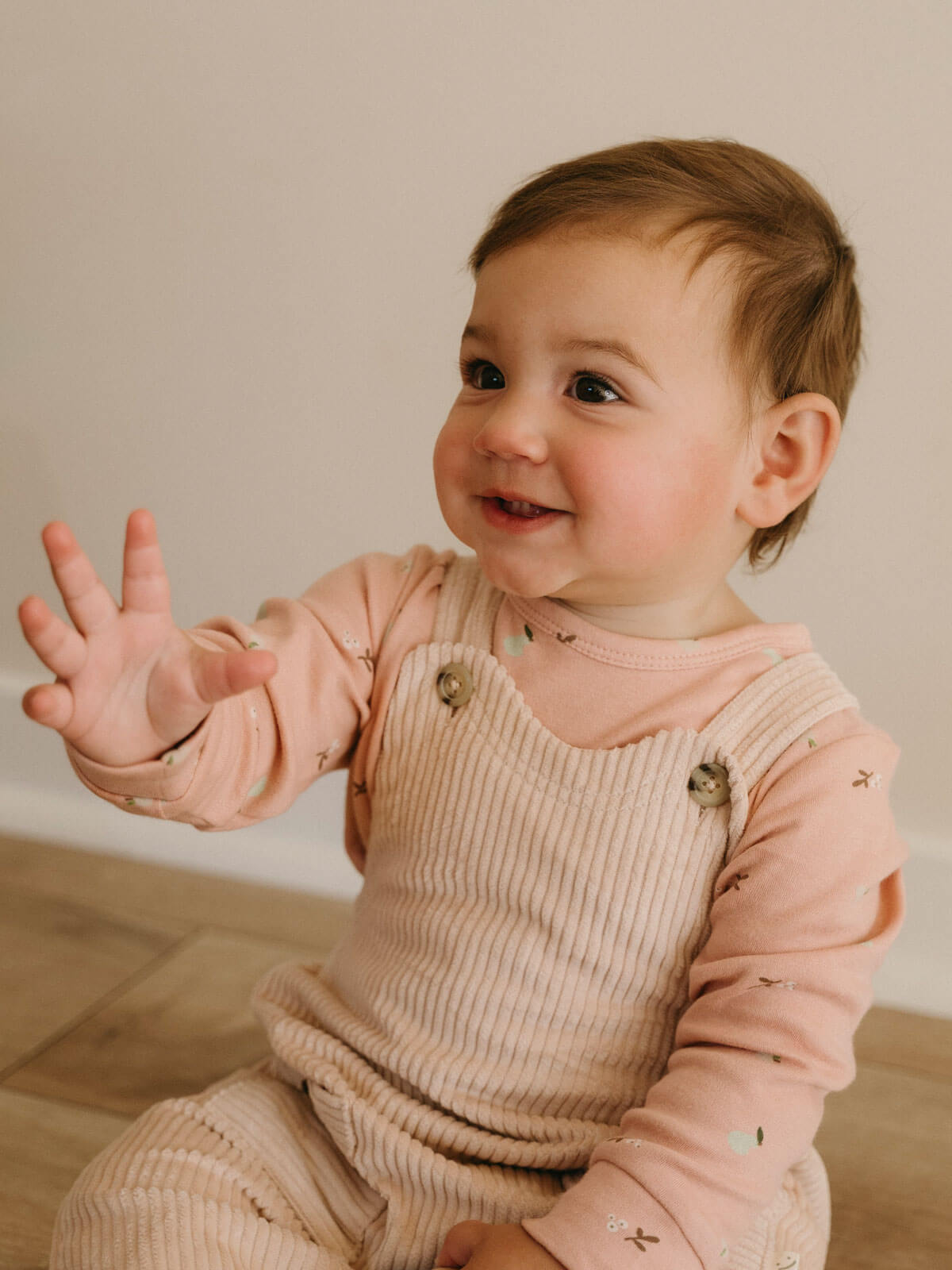 Baby wearing a pink long-sleeve shirt and beige overalls, sitting on a wooden floor.