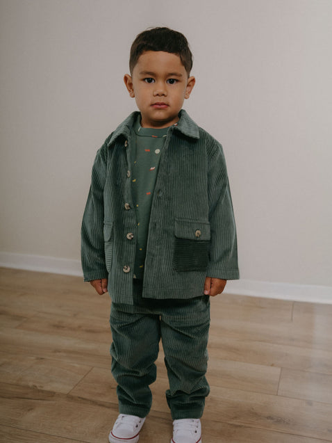 Child wearing a green corduroy outfit standing on a wooden floor. Corduroy Pants - Balsam Girls and Boys