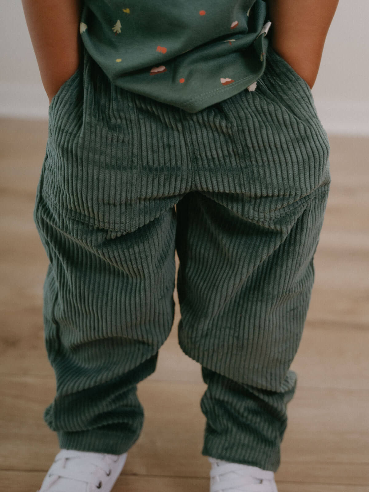 Boy wearing green corduroy pants and a matching top on a wooden floor. Corduroy Pants - Balsam Girls and Boys
