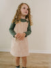 Young girl wearing a green long-sleeve shirt and beige pinafore dress standing on a wooden floor. Corduroy Pinafore Dress - Clay Girls
