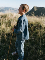 Young person in denim outfit standing in grassy field with mountains behind Denim Pants - Blue Denim Girls and Boys