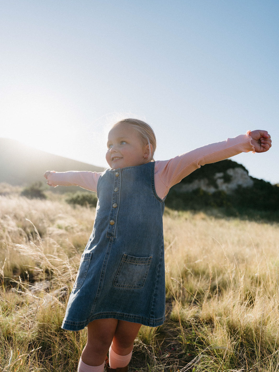 Young girl in denim pinafore and pink shirt standing in grassy field with arms outstretched Denim Pinafore Dress - Blue Denim Girls