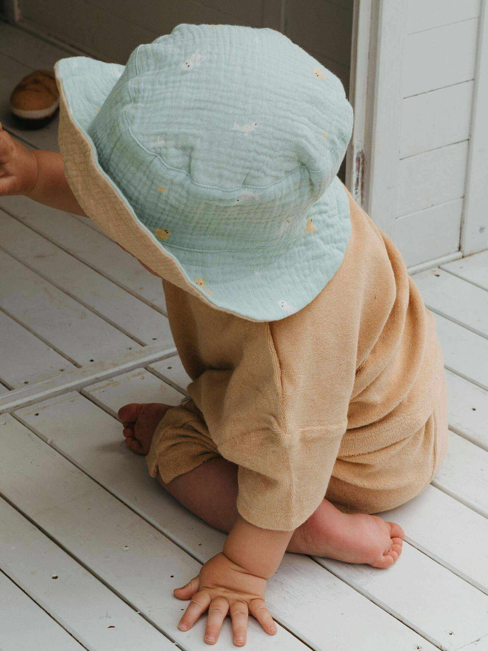 Child wearing a light blue bucket hat and beige outfit on a white wooden floor.