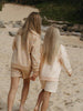 Two children in matching beige outfits walking on a sandy beach.
