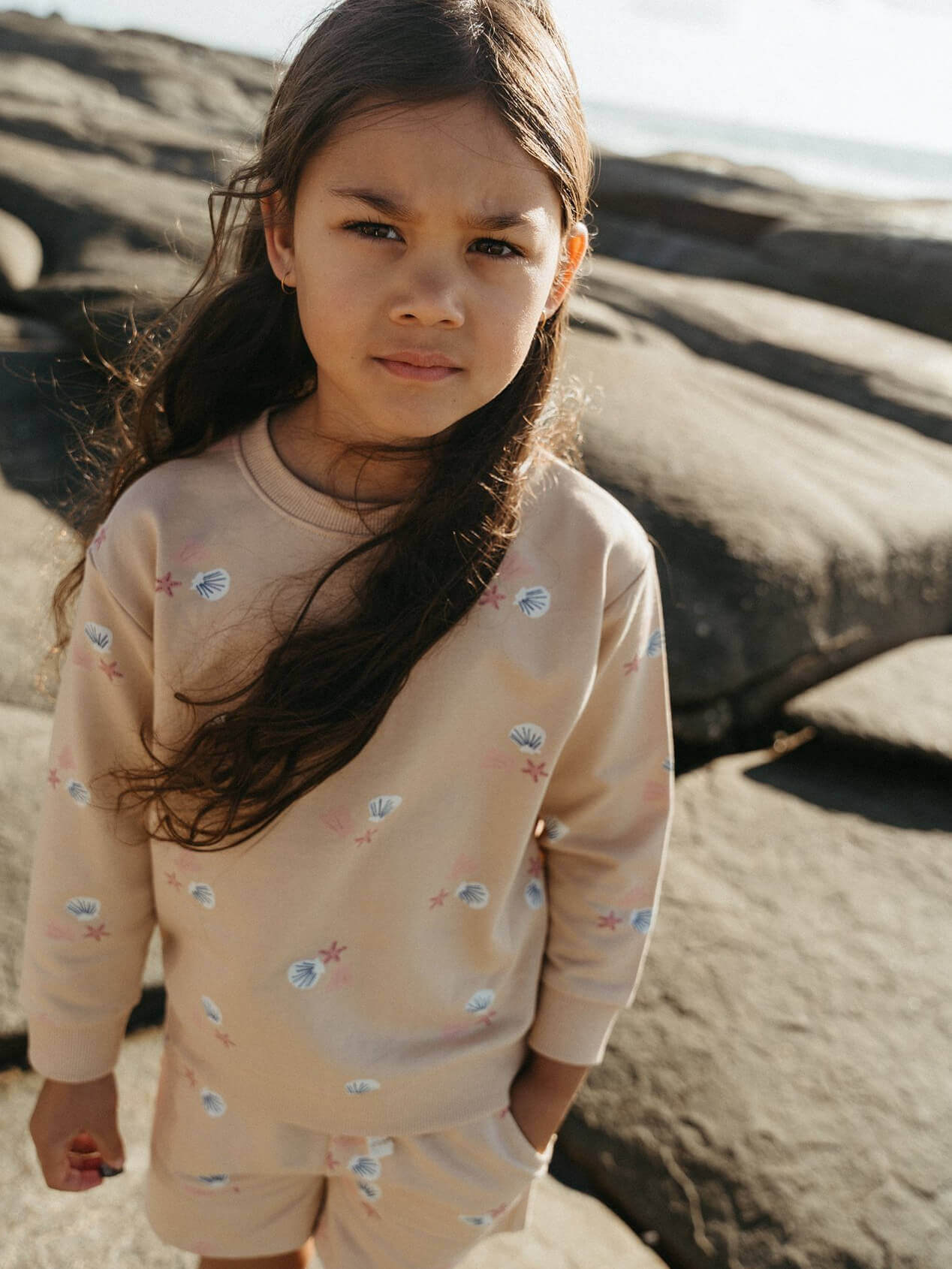 Young girl in a patterned outfit standing on a sandy beach.