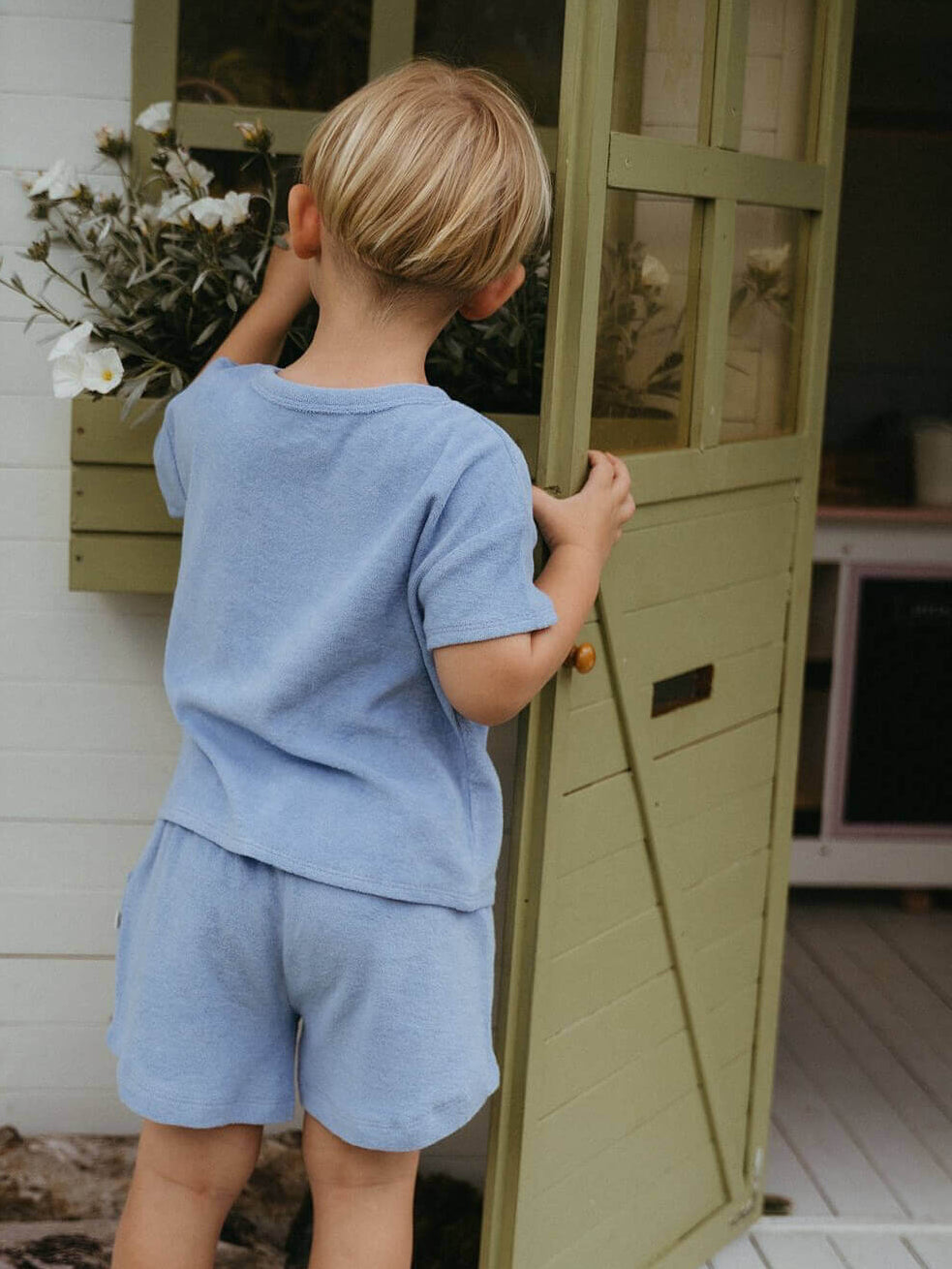 Child in a blue outfit standing in front of a green door on a wooden deck.