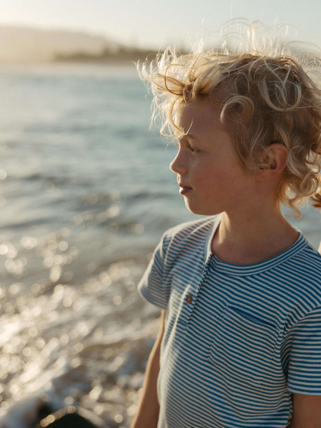 A young child with curly blond hair stands by the water at sunset, wearing a blue and white striped short-sleeve shirt.