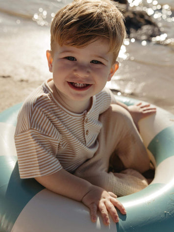Toddler boy smiling to the camera sitting on a back float wearing a neutral striped baby tee 