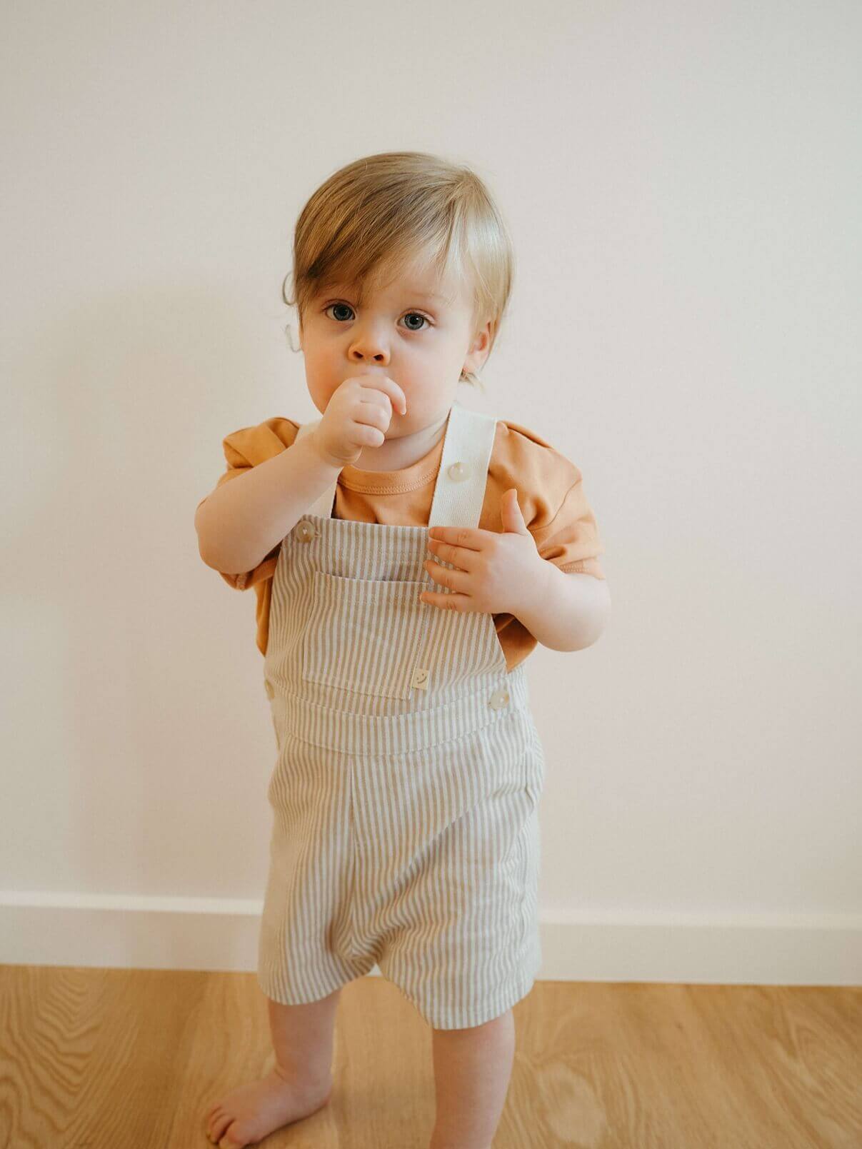 Child wearing a striped romper and orange shirt against a plain wall.