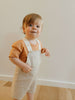 Child wearing a brown shirt and white striped overalls standing against a plain wall.