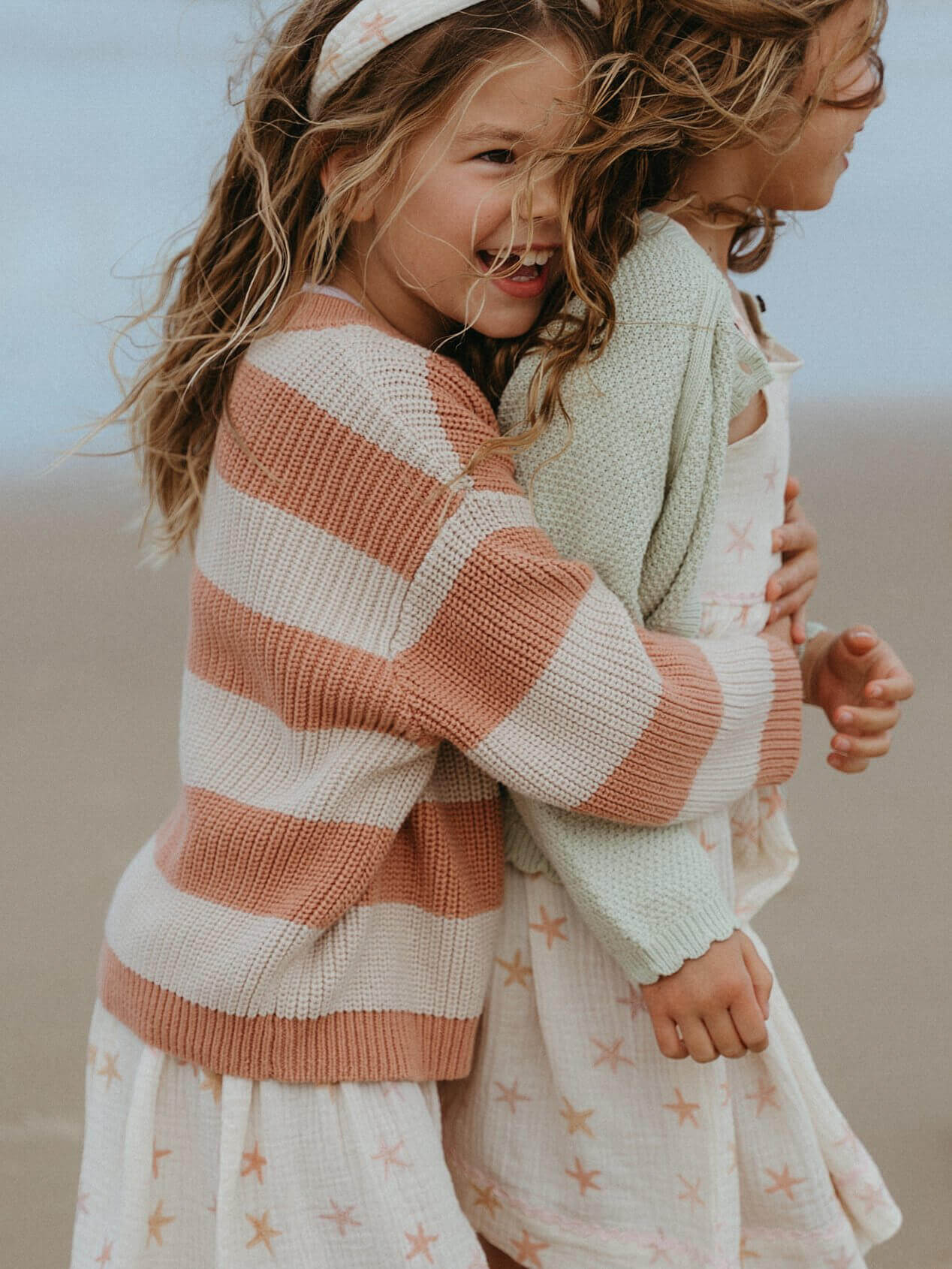 Two children hugging, one wearing a striped sweater and the other in a star-patterned dress, against a neutral background.