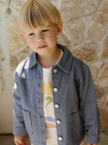 Child wearing a denim jacket over a colorful shirt against a stone wall background