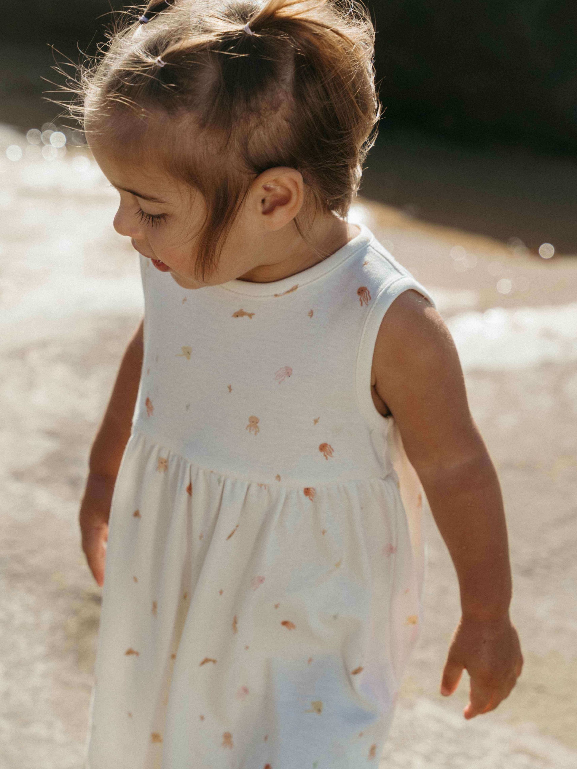 Child in a white dress with star pattern standing on a sandy beach.