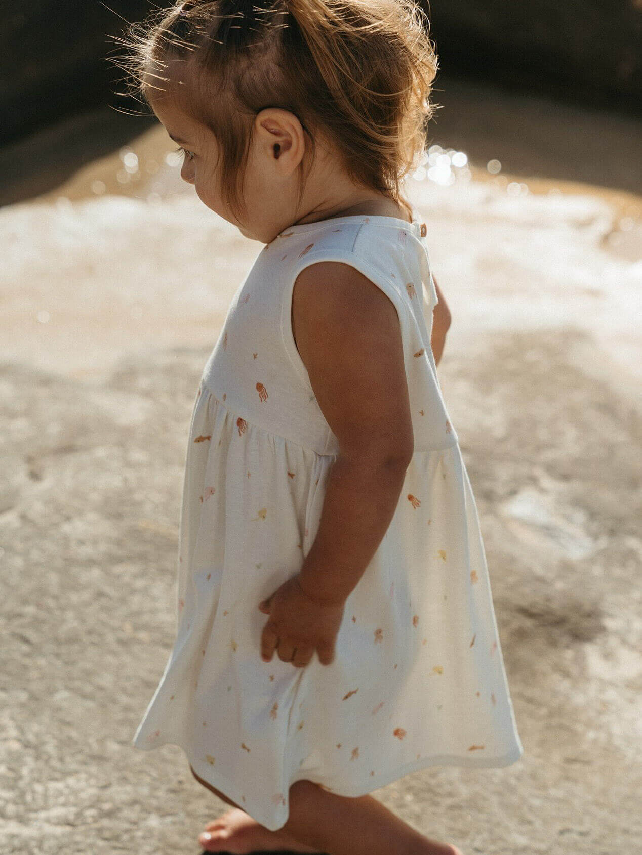 Child in a white dress with colorful patterns standing on a sandy beach.