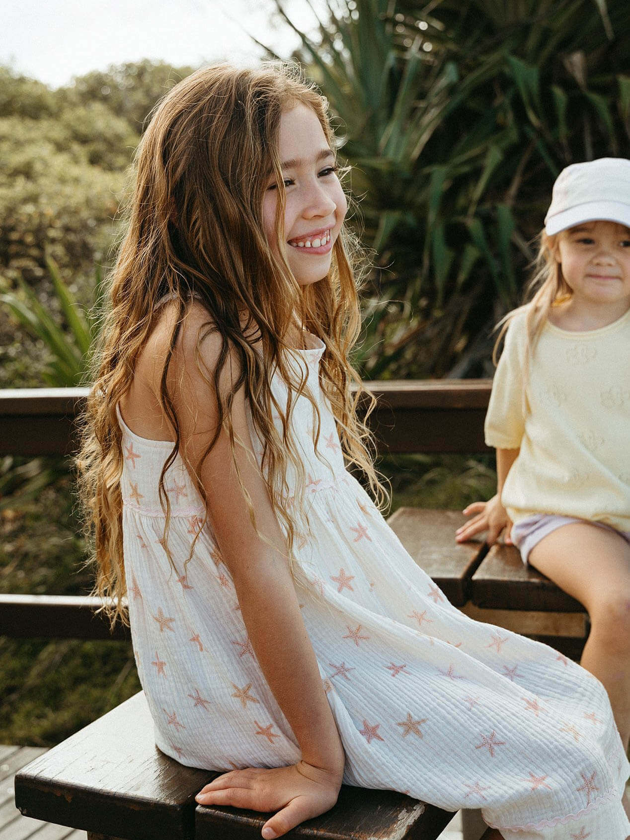 Two children sitting on a wooden bench outdoors with greenery in the background