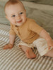 Baby sitting on a striped surface wearing a brown top and beige pants.