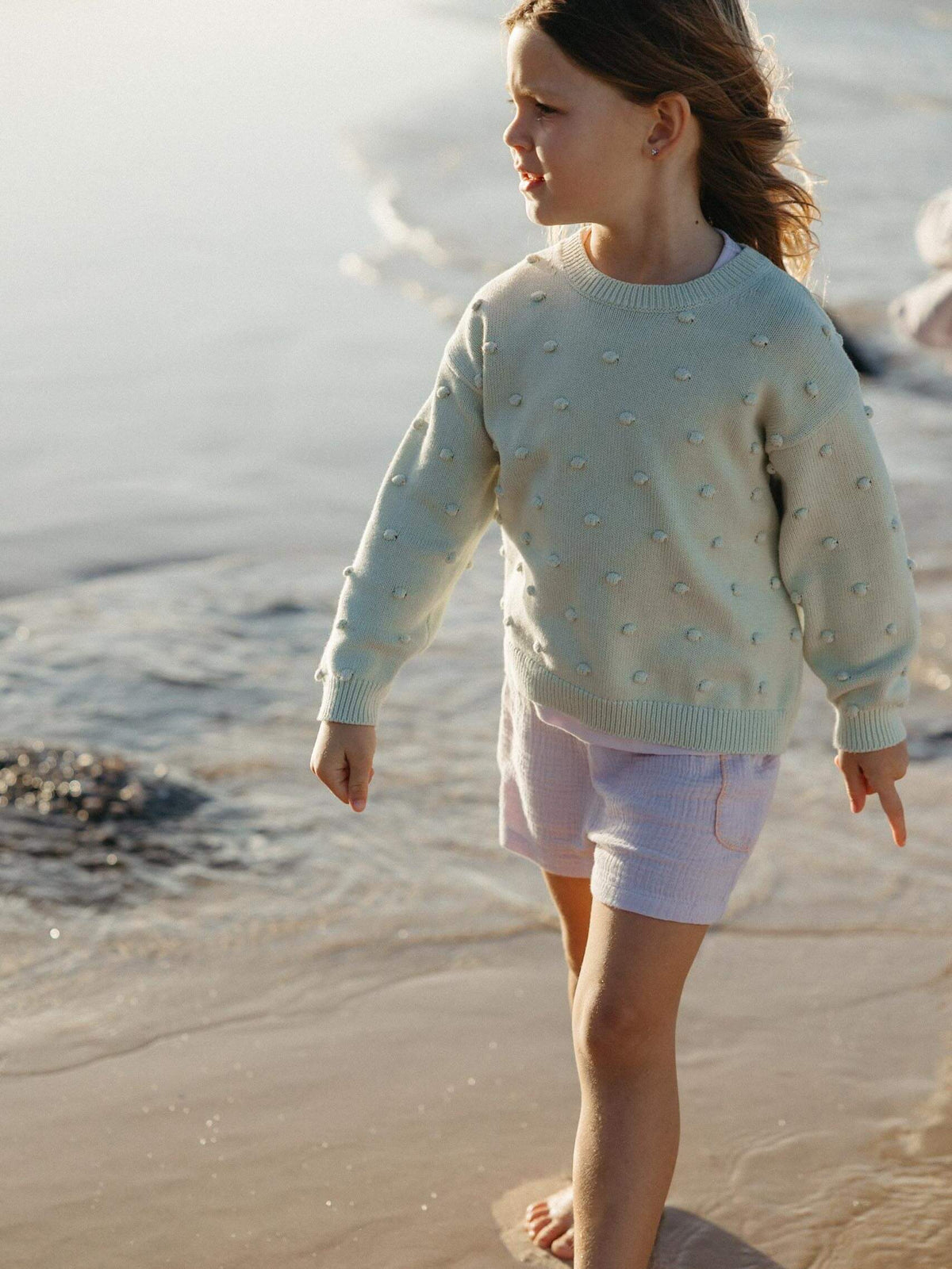A young girl walking barefoot on a sandy beach near the water, wearing a pale mint green knit sweater with raised pearl-like details and white shorts.