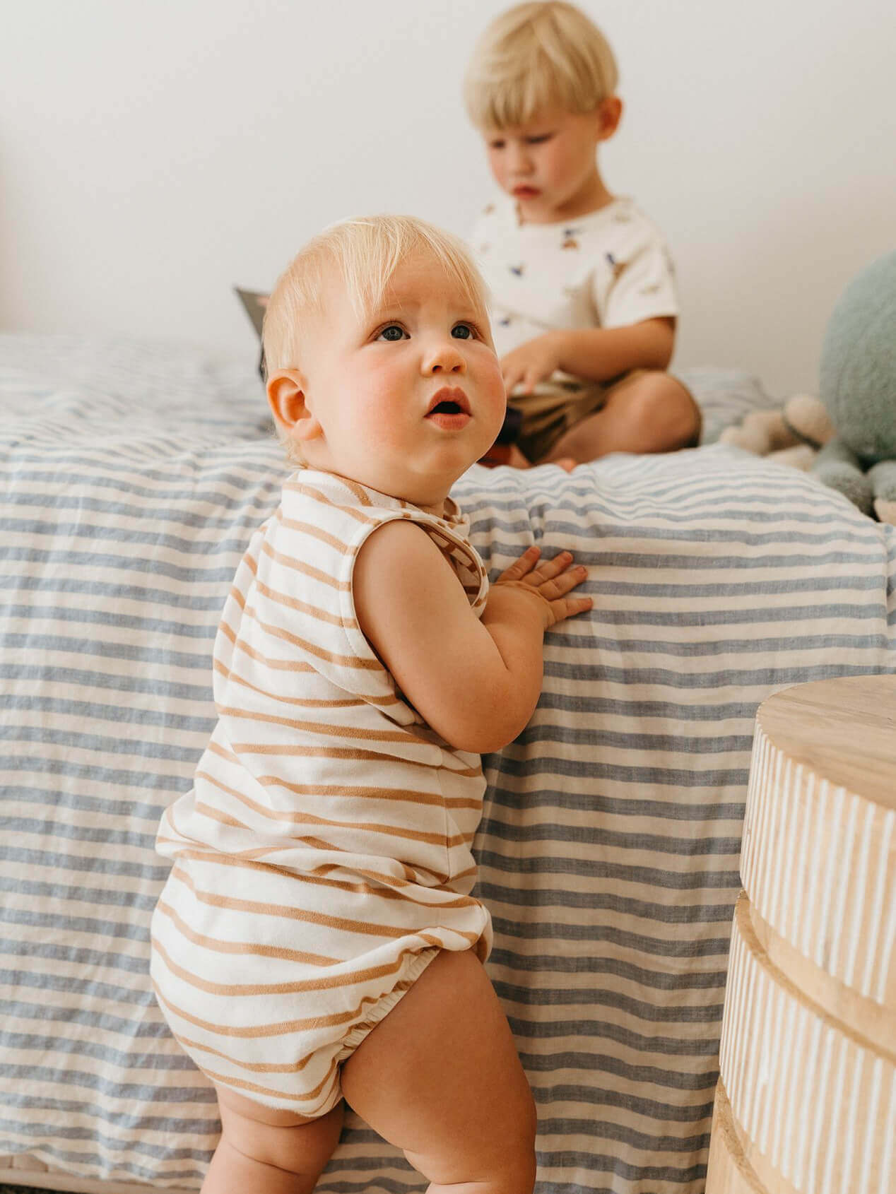 Baby in a striped romper standing on a bed with a toddler in the background.