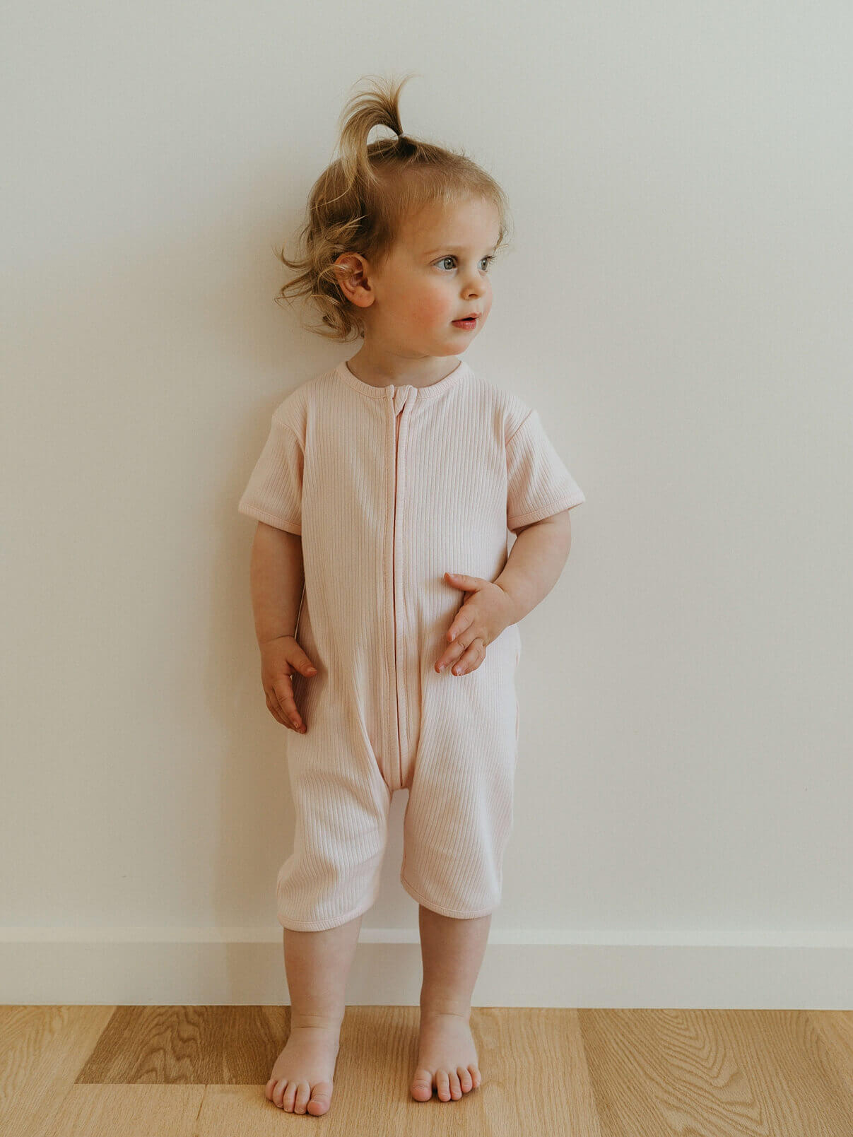 Child wearing a light pink onesie standing against a plain wall.