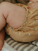 Close-up of a baby wearing a mustard yellow bloomers with floral patterns on a striped fabric background.