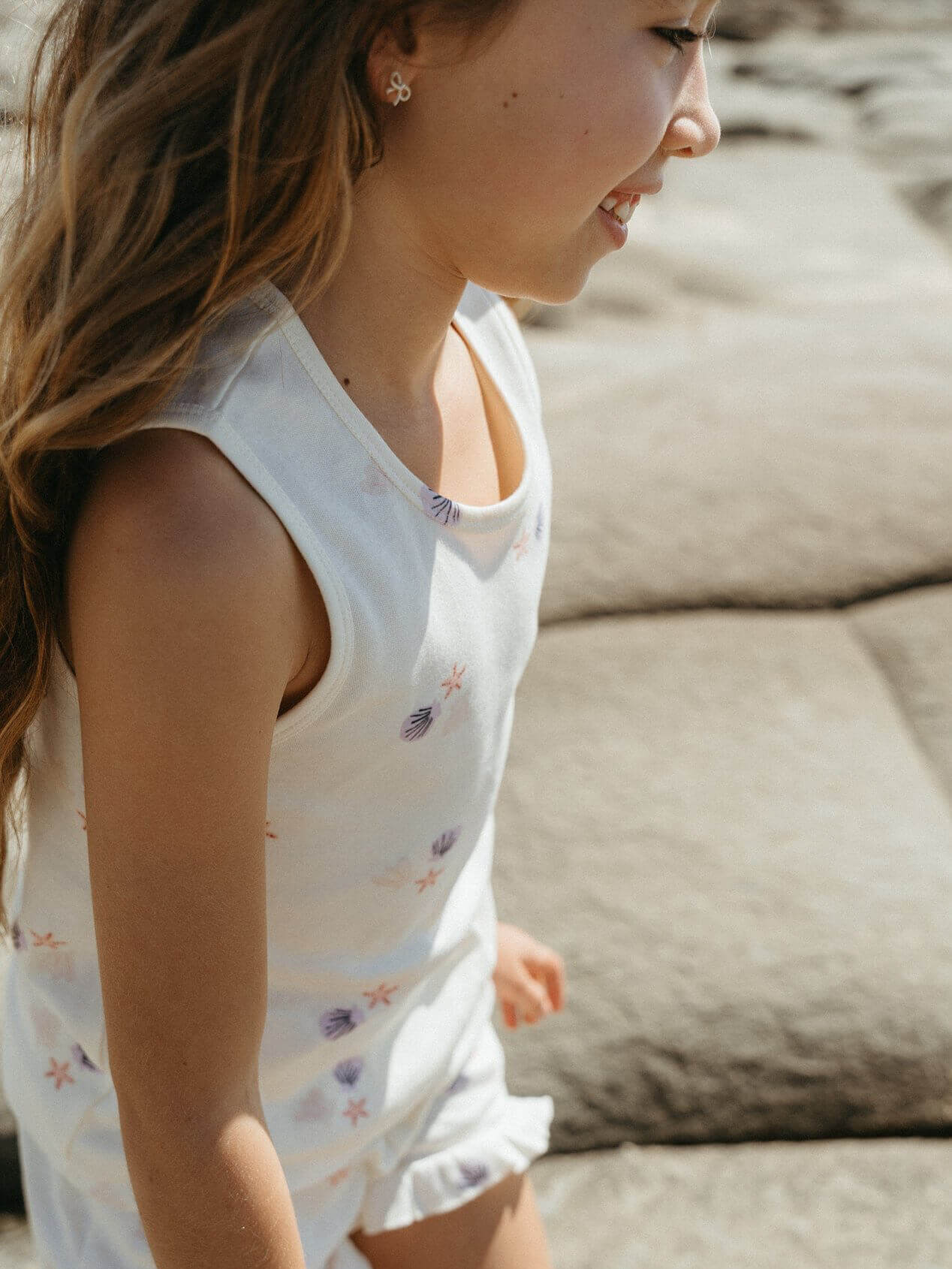 Young girl in a white sleeveless dress with floral patterns sitting on a stone surface.