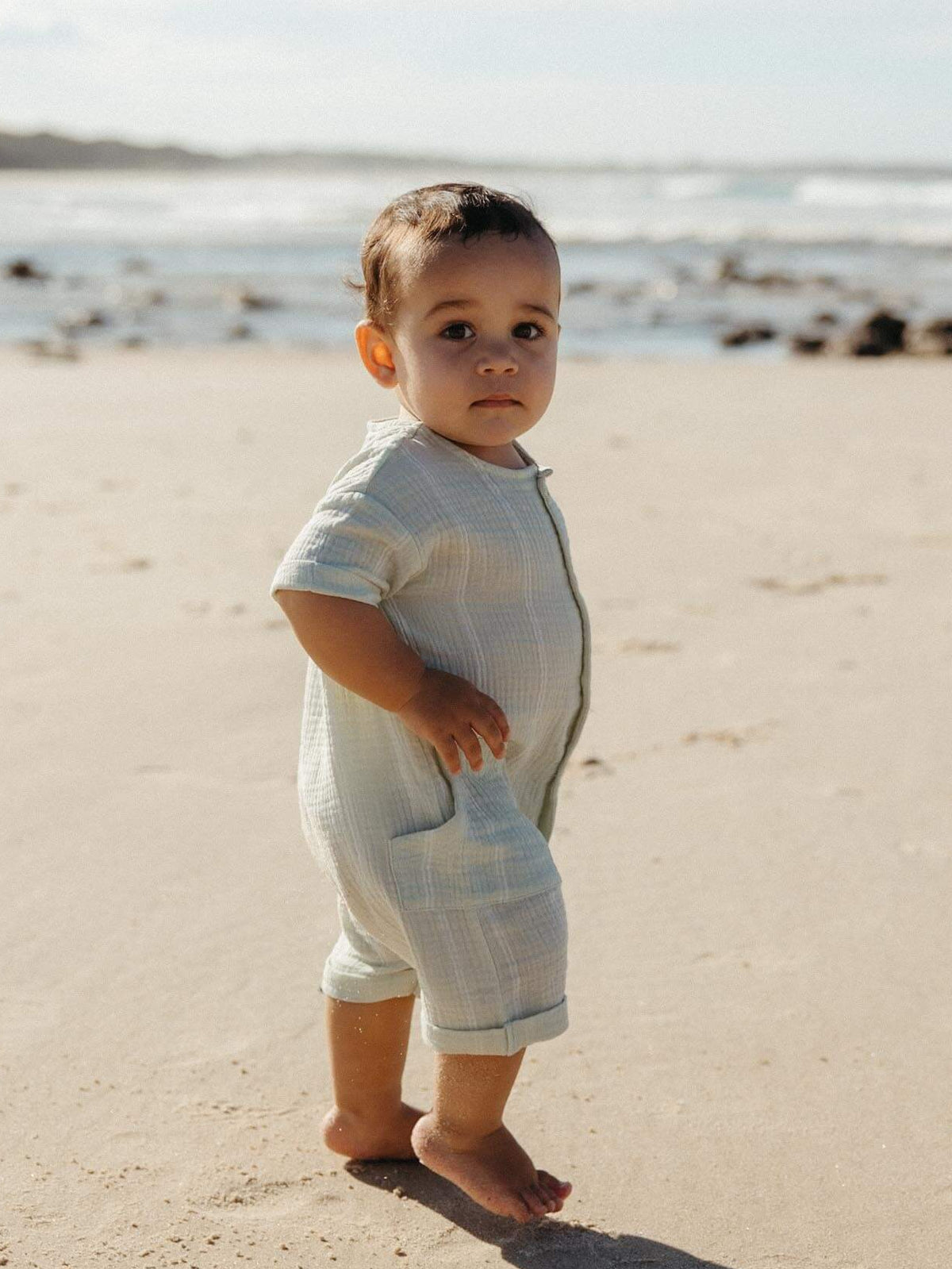 little boy walking on the sand wearing a light green striped romper