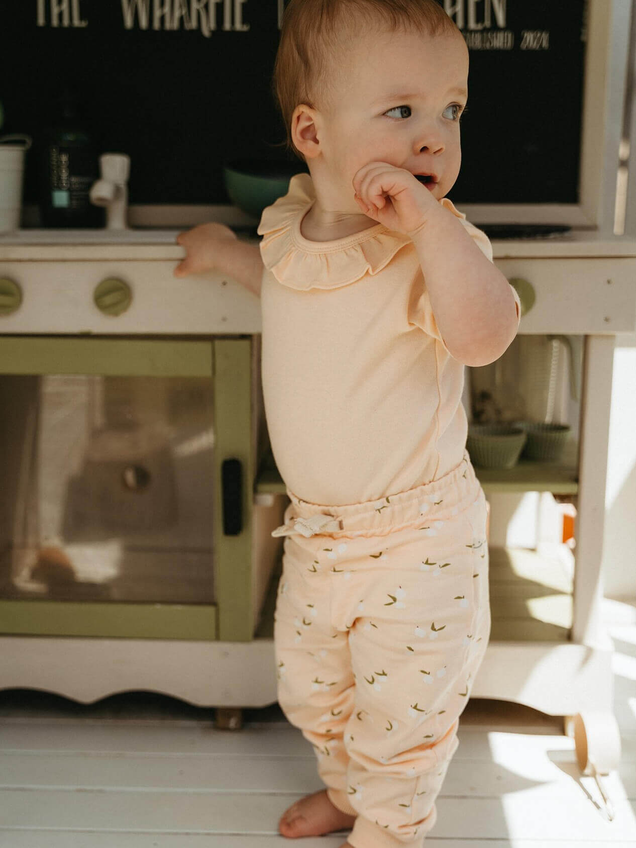 Baby in a peach outfit standing in a kitchen