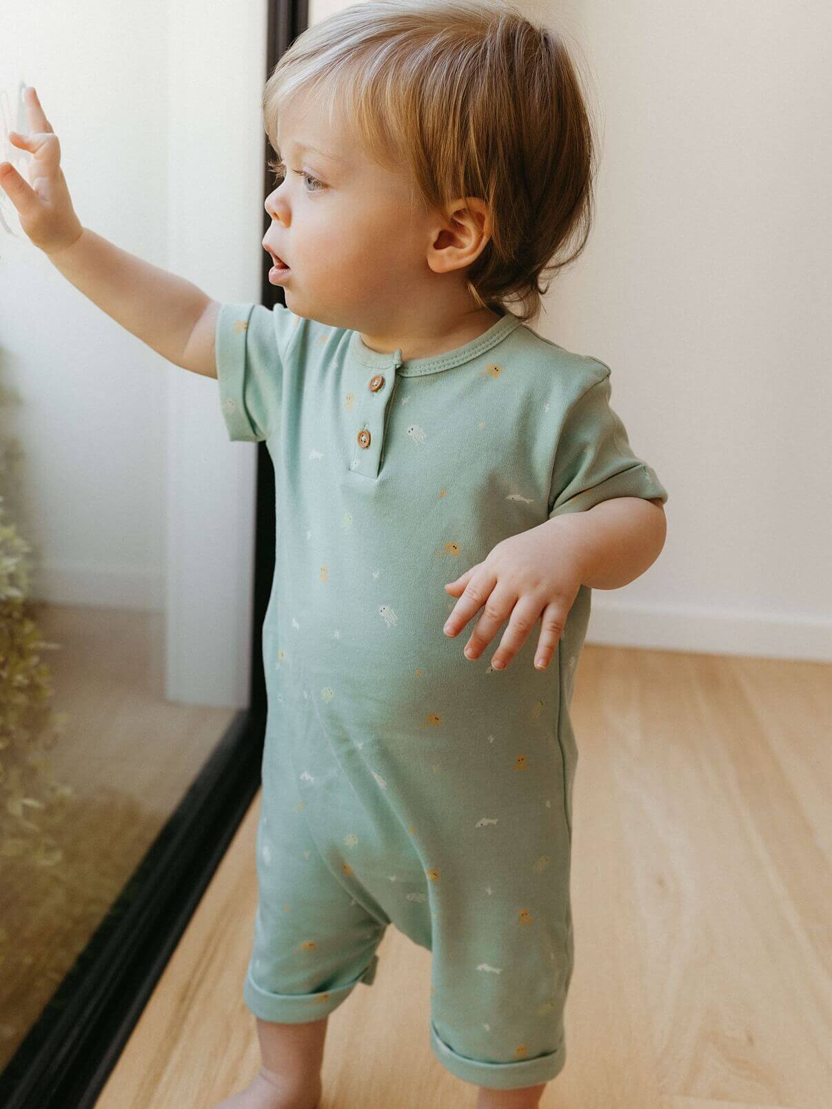Child wearing a light green onesie with small patterns, standing indoors.