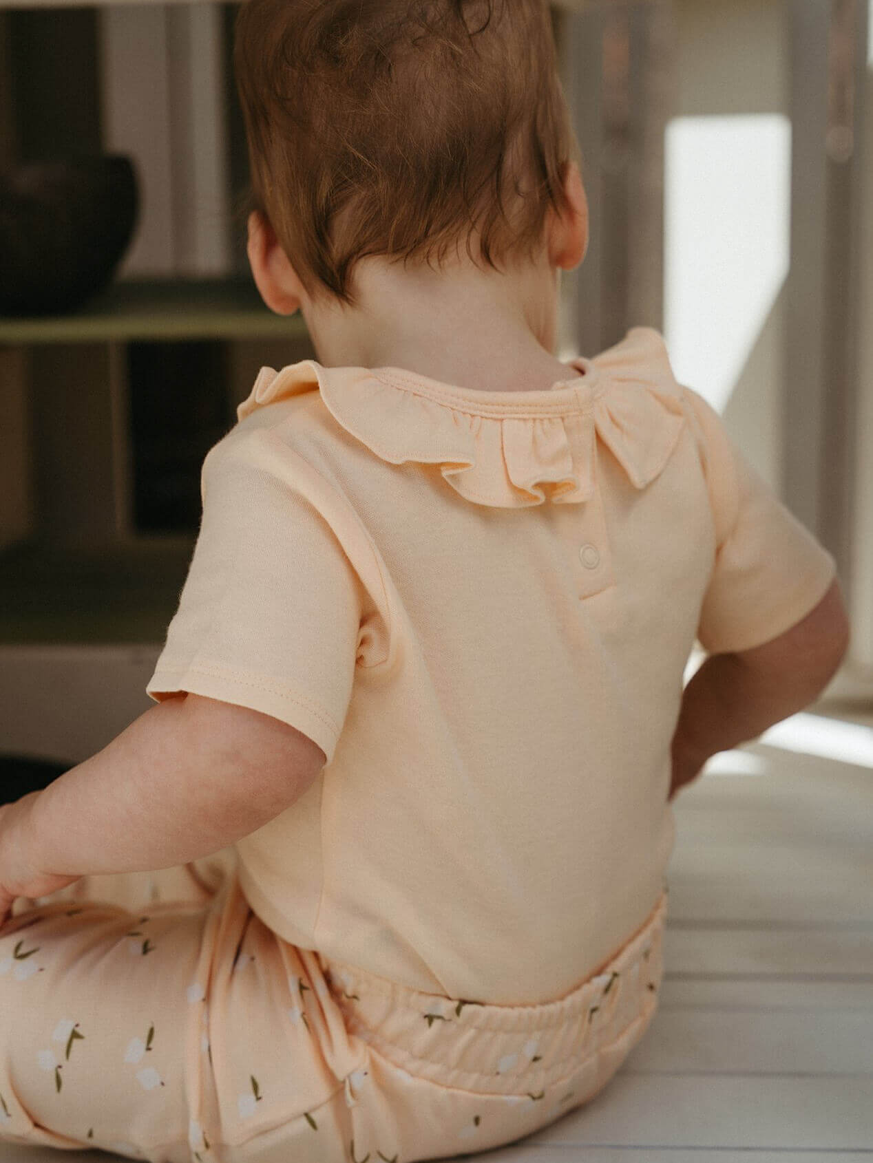 Baby wearing a light-colored outfit with ruffled collar sitting on a surface.