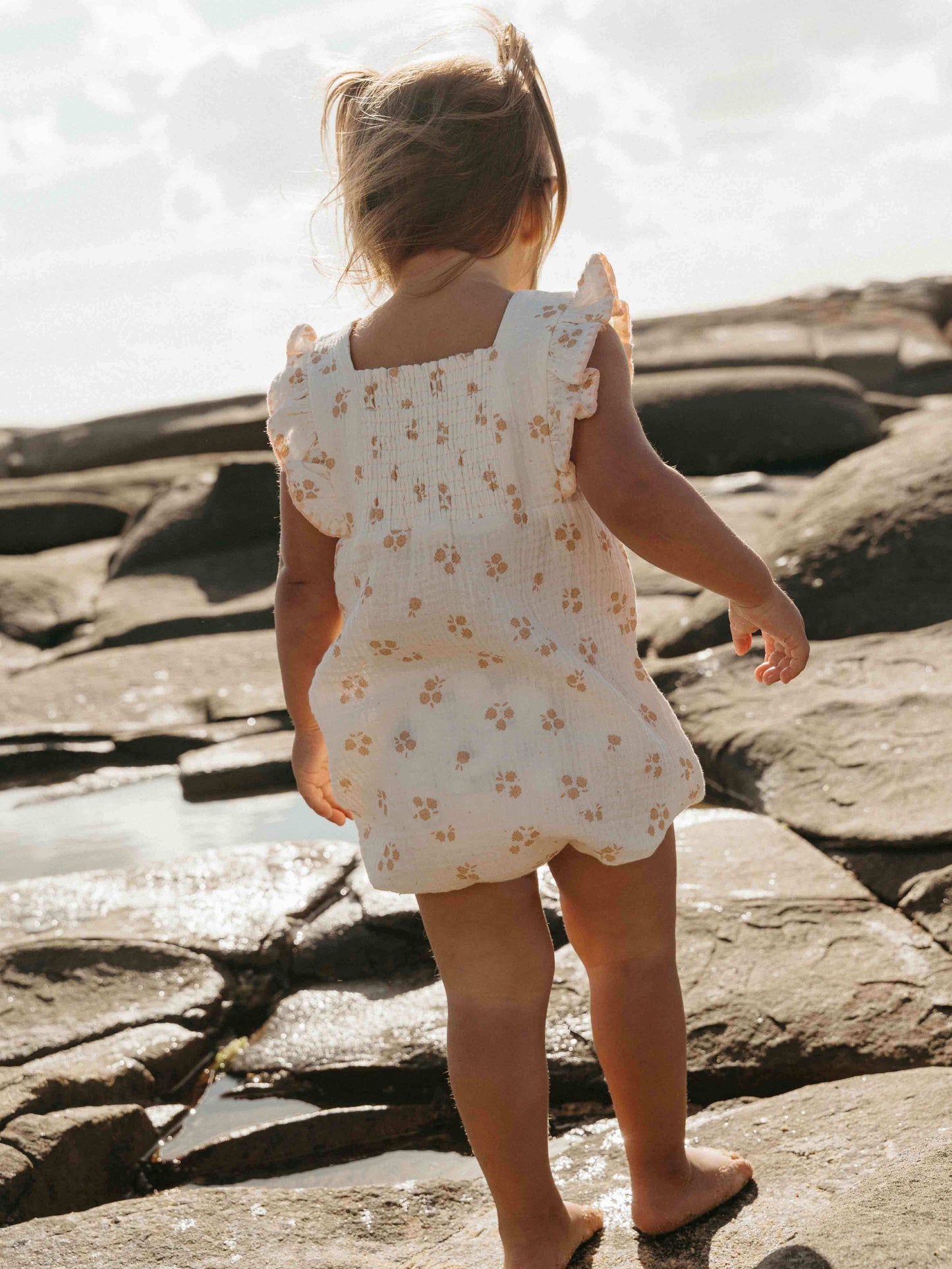 Child in a floral romper standing on rocky terrain near water
