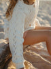 A close-up lifestyle shot of a child wearing a cream-colored pointelle cardigan and peach shorts, sitting on a log in a sandy beach setting.