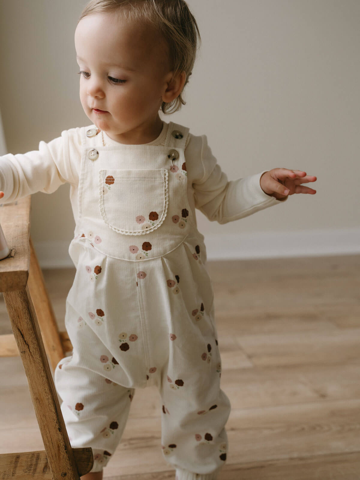 Toddler wearing cream overalls with floral print standing by wooden furniture Printed Overalls - Autumn Bouquet Ecru Baby girl