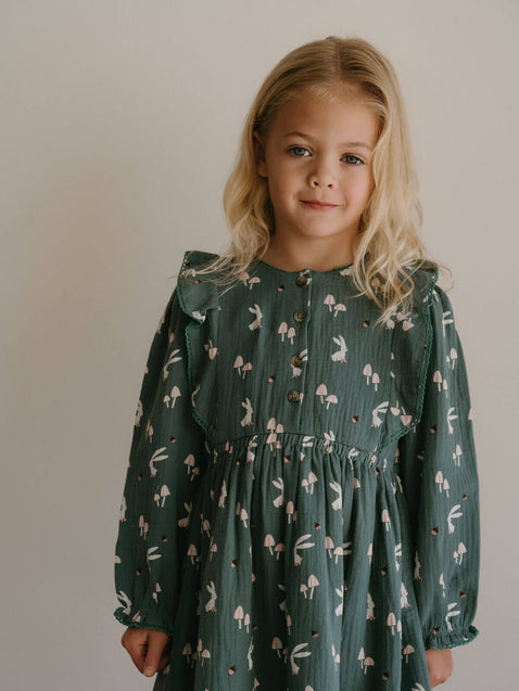 Young girl wearing a green dress with a pattern of small figures against a plain background Printed Woven Dress - Forest Gathering Balsam Girls