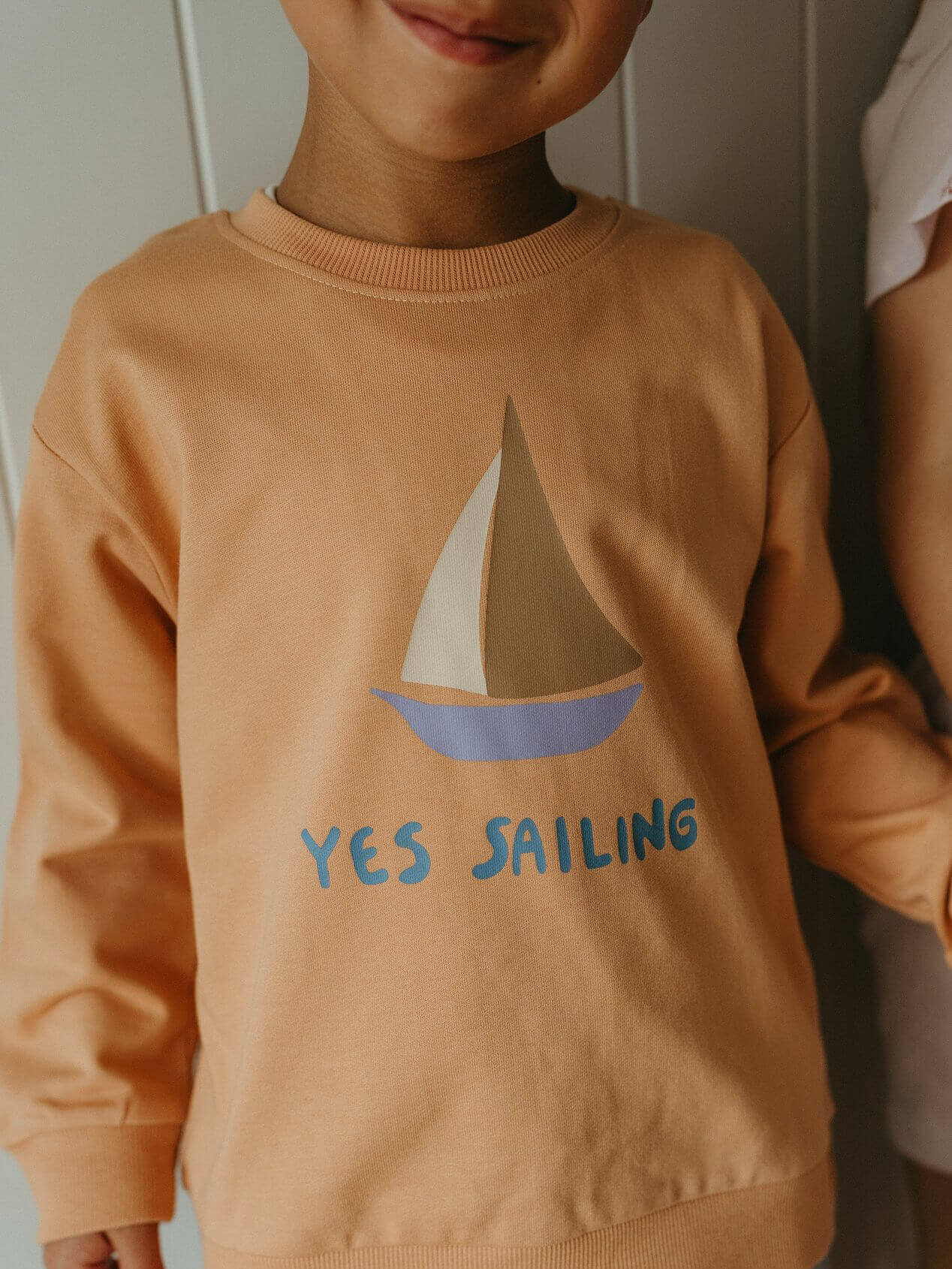 Child wearing a tan sweatshirt with a sailboat design and 'YES SAILING' text.