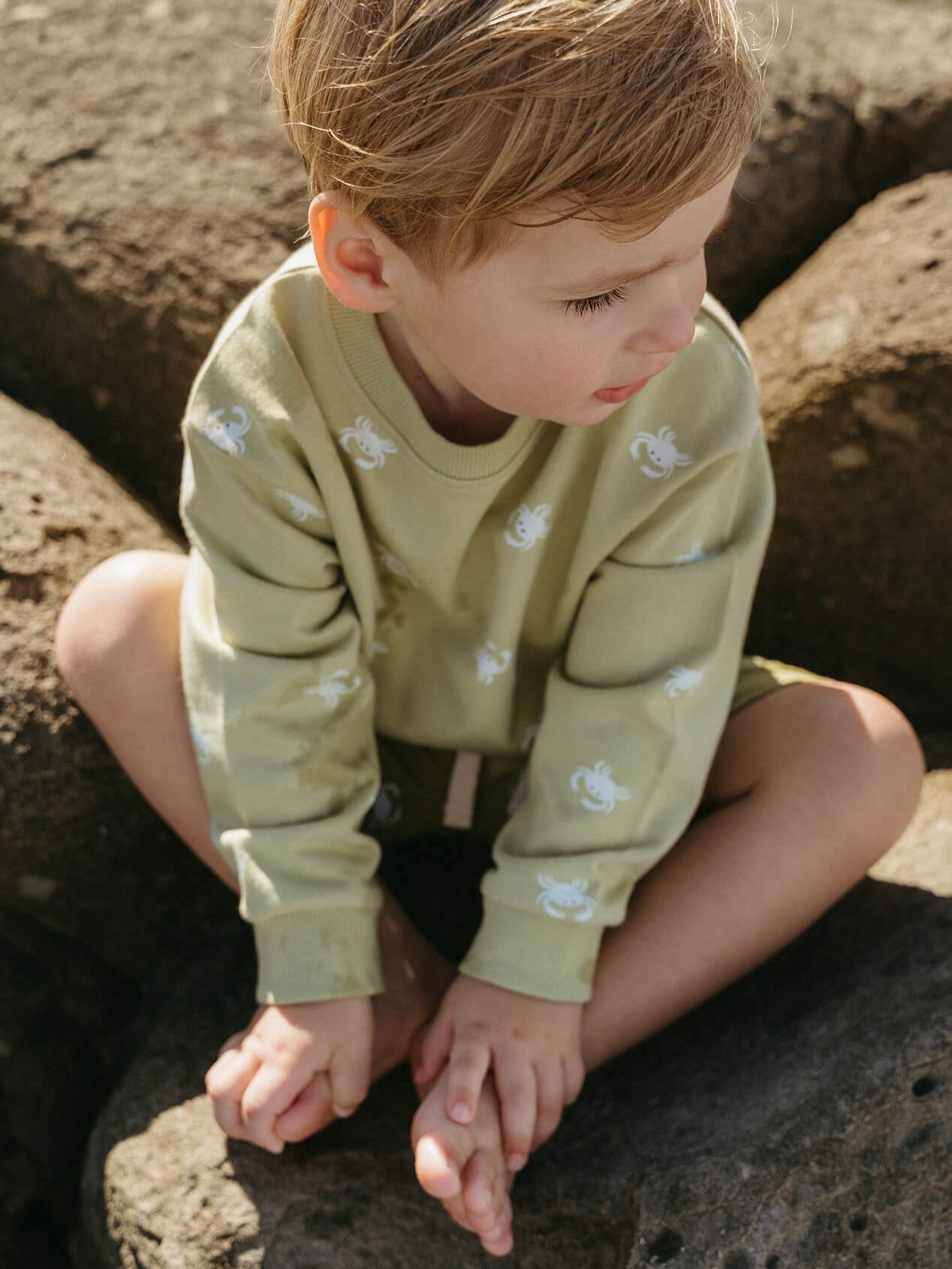 Child wearing a green sweater with floral patterns sitting on rocks.