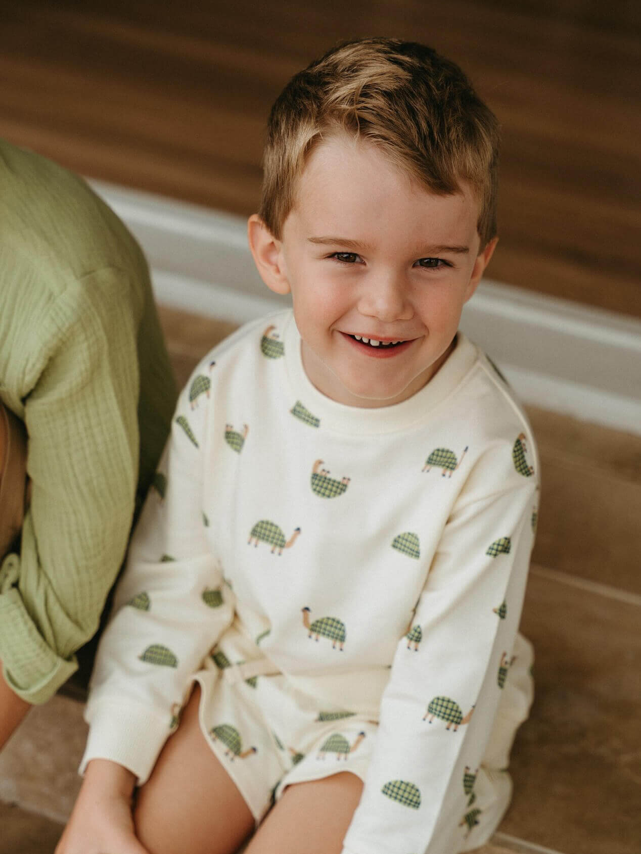 Child wearing a white sweatshirt with green animal patterns, sitting on a wooden floor.