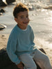 A smiling young boy sitting on a rock by the water, wearing a light blue knit sweater and striped pants.