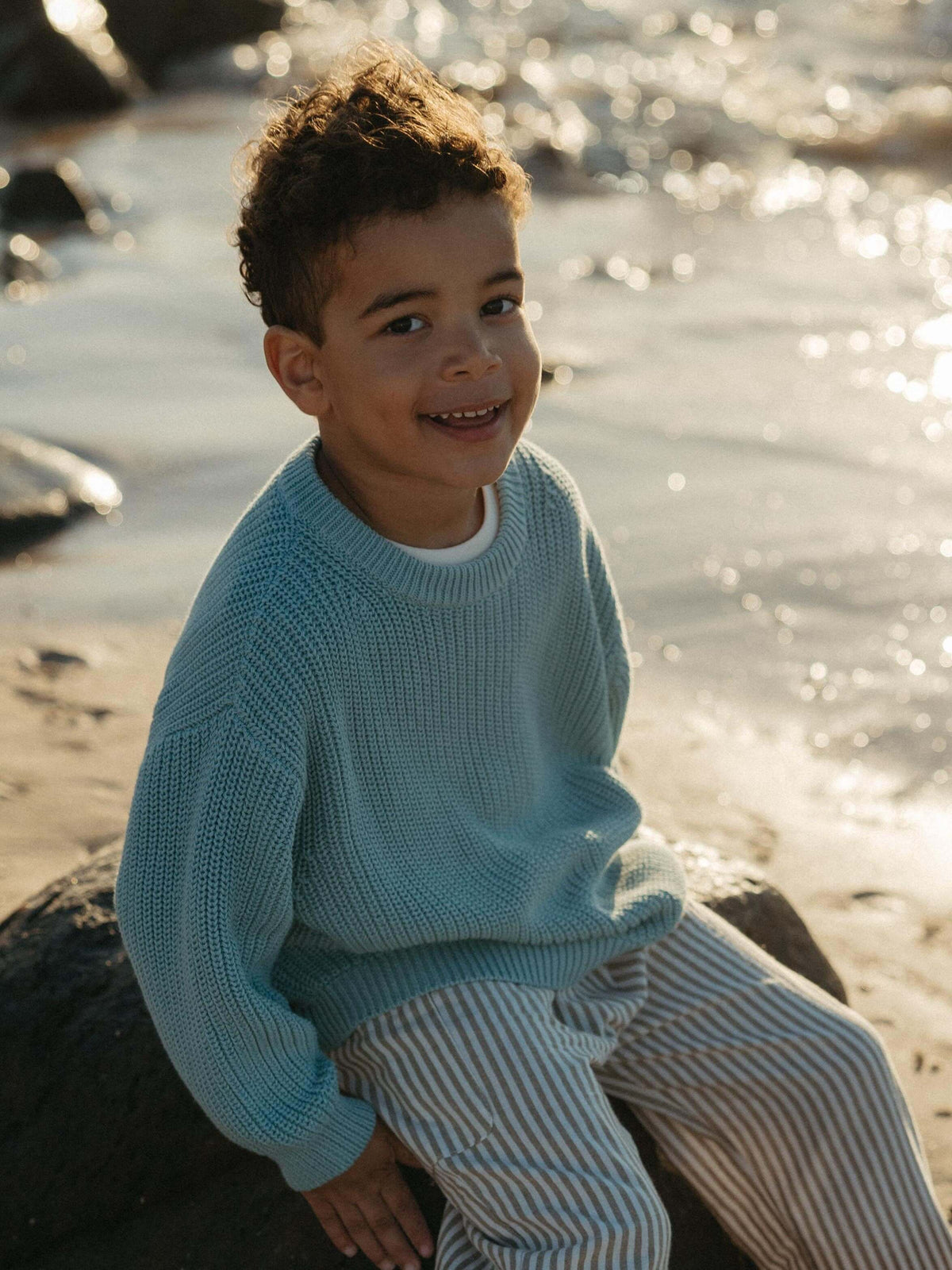 A smiling young boy sitting on a rock by the water, wearing a light blue knit sweater and striped pants.