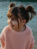 A young girl with brown hair in two buns wearing a pink ribbed knit sweater, smiling and looking to the side against a blurred outdoor background.