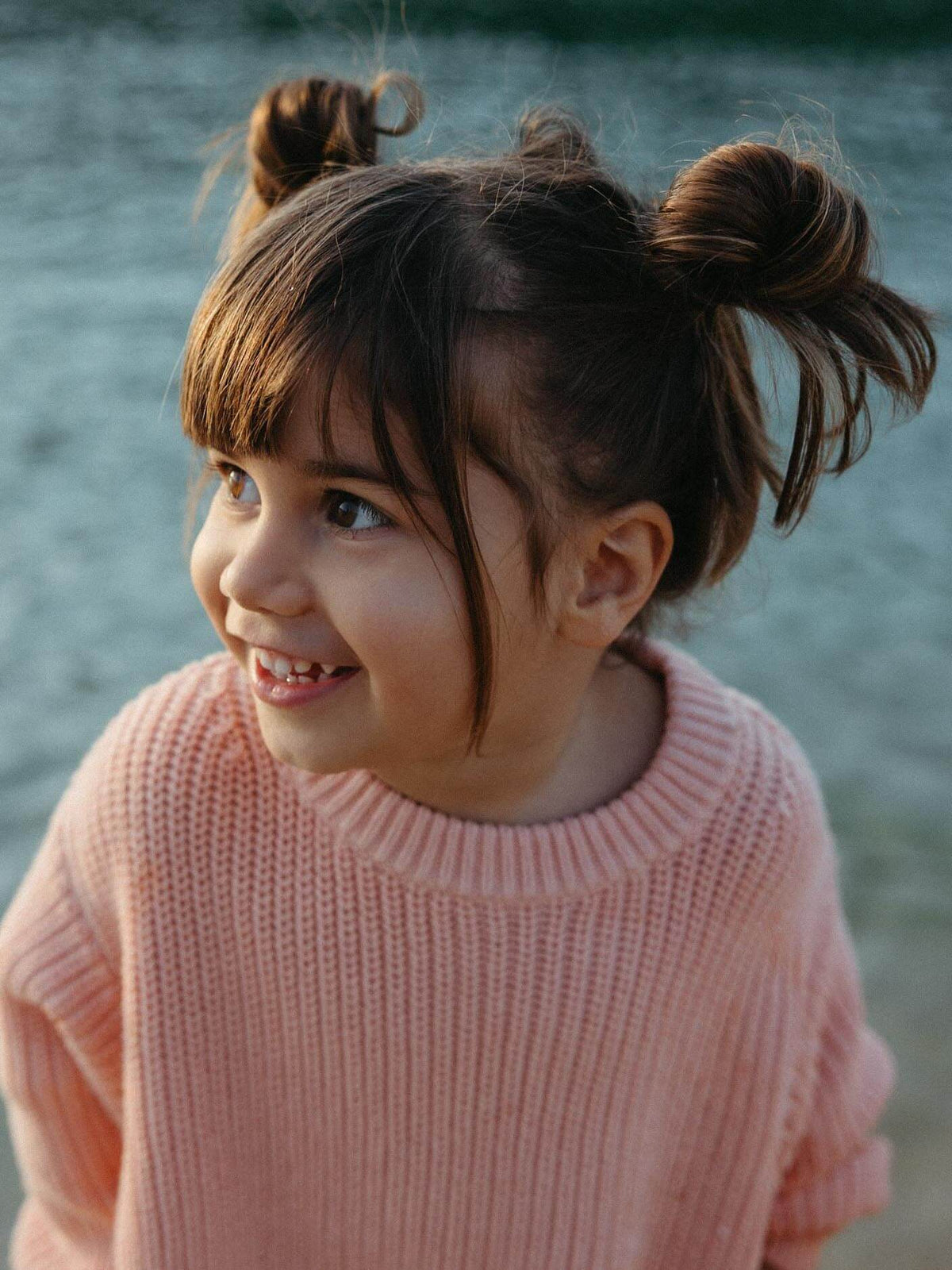 A young girl with brown hair in two buns wearing a pink ribbed knit sweater, smiling and looking to the side against a blurred outdoor background.
