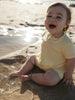 A baby sitting on wet sand at the beach wearing a pale yellow short-sleeve romper, looking up and smiling with the sun reflecting on the water behind them.