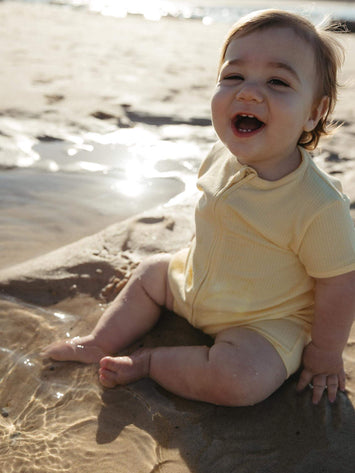A baby sitting on wet sand at the beach wearing a pale yellow short-sleeve romper, looking up and smiling with the sun reflecting on the water behind them.