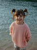 A young girl with brown pigtails smiles while standing in shallow water, wearing a soft pink ribbed knit sweater and striped pants.