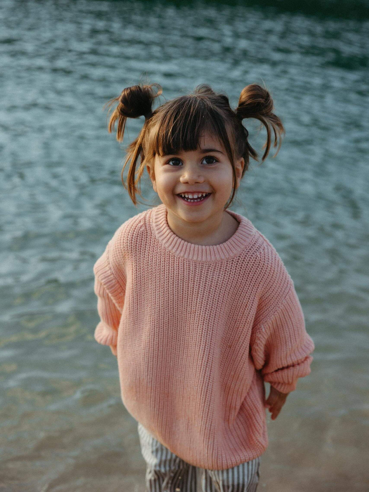 A young girl with brown pigtails smiles while standing in shallow water, wearing a soft pink ribbed knit sweater and striped pants.