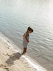 A young child stands in shallow water at the shoreline, wearing a sleeveless light blue and white vertically striped dress. The background is a natural outdoor setting with calm water and wet sand.